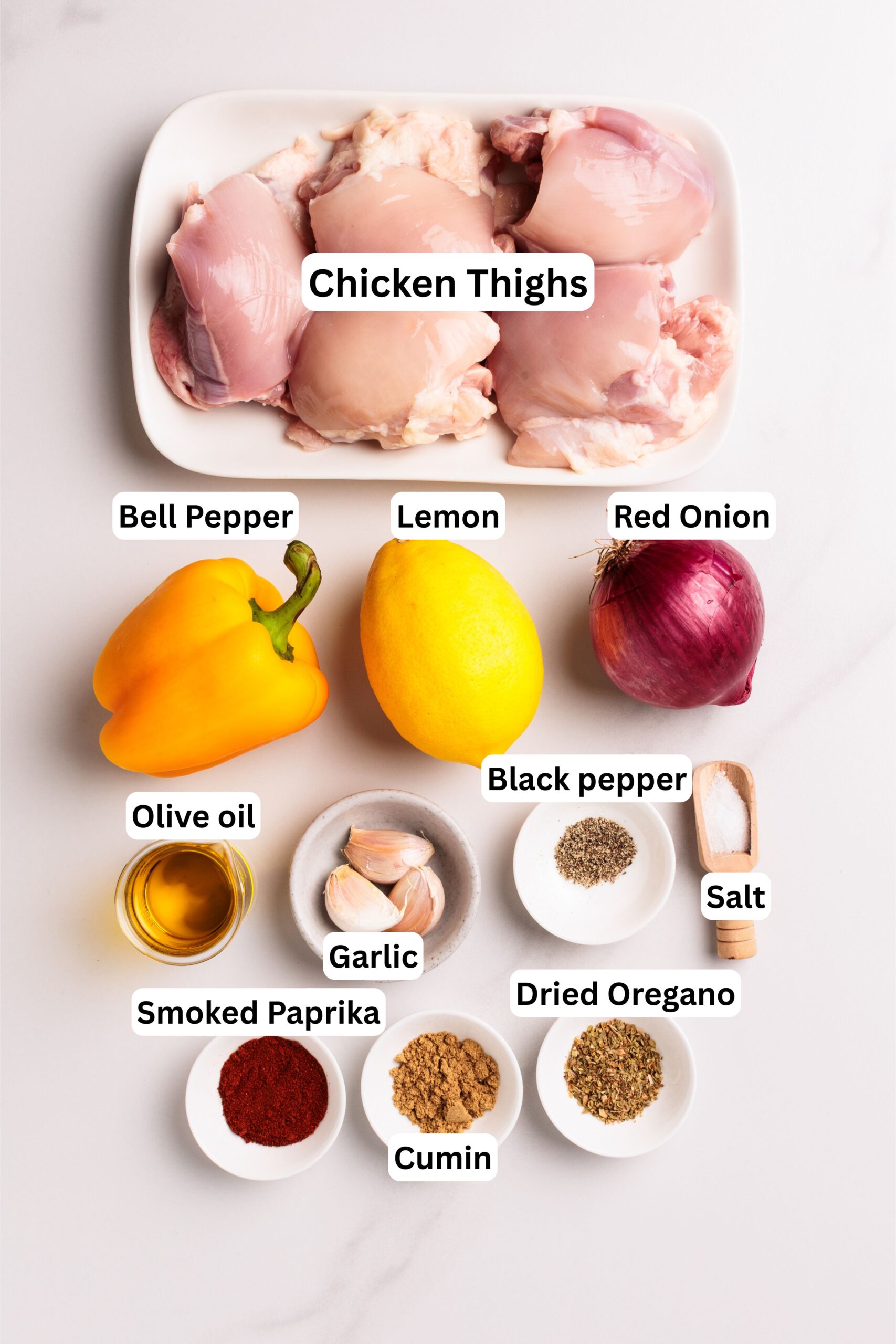 Overhead view of labeled ingredients on a white surface: chicken thighs, yellow bell pepper, lemon, red onion, olive oil, garlic, black pepper, salt, smoked paprika, cumin, and dried oregano in small bowls.