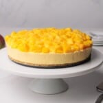 A no-bake cheesecake topped with diced mango pieces sits on a white cake stand.