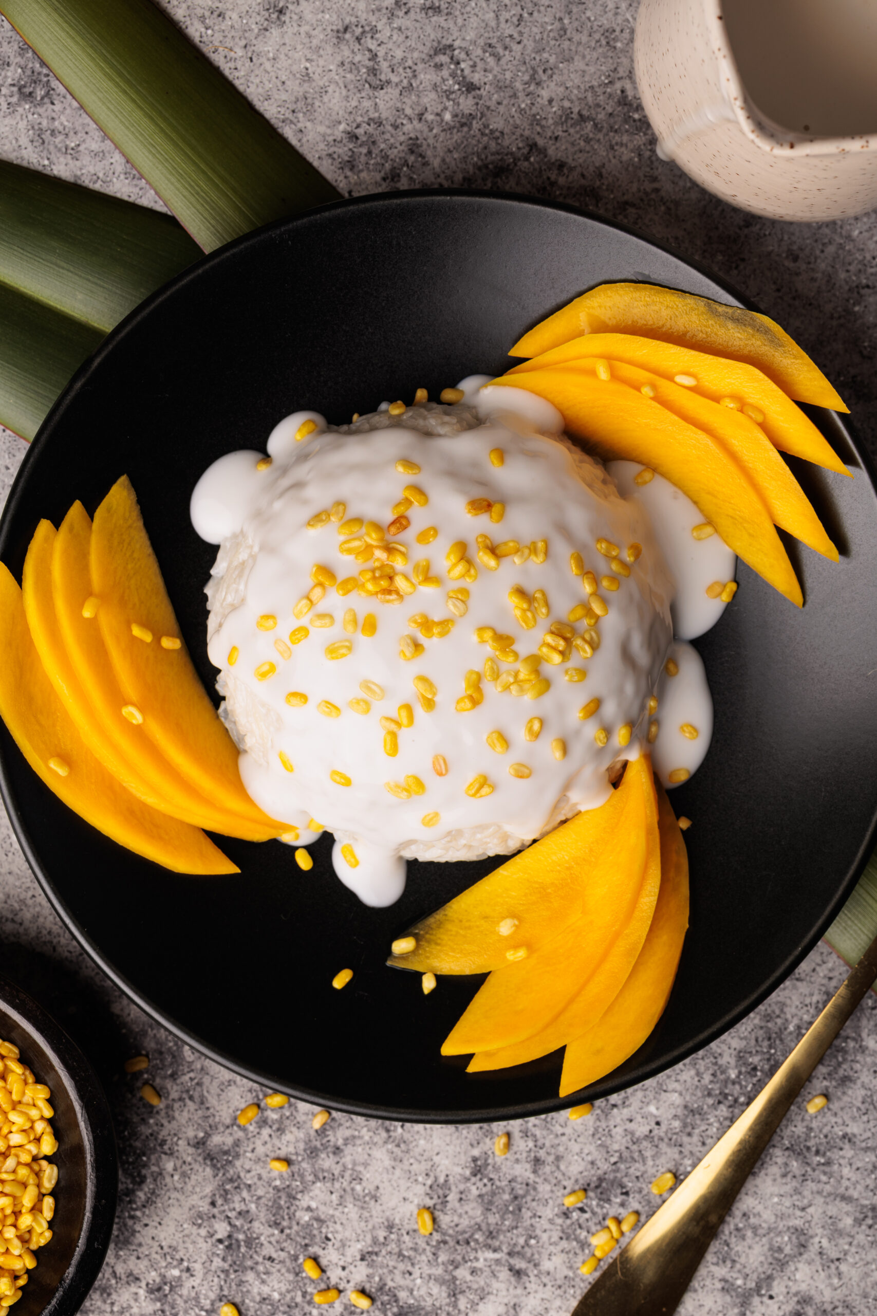 A black plate with sticky rice topped with coconut cream and yellow mung beans, surrounded by neatly arranged mango slices. A cup and green leaves are in the background.