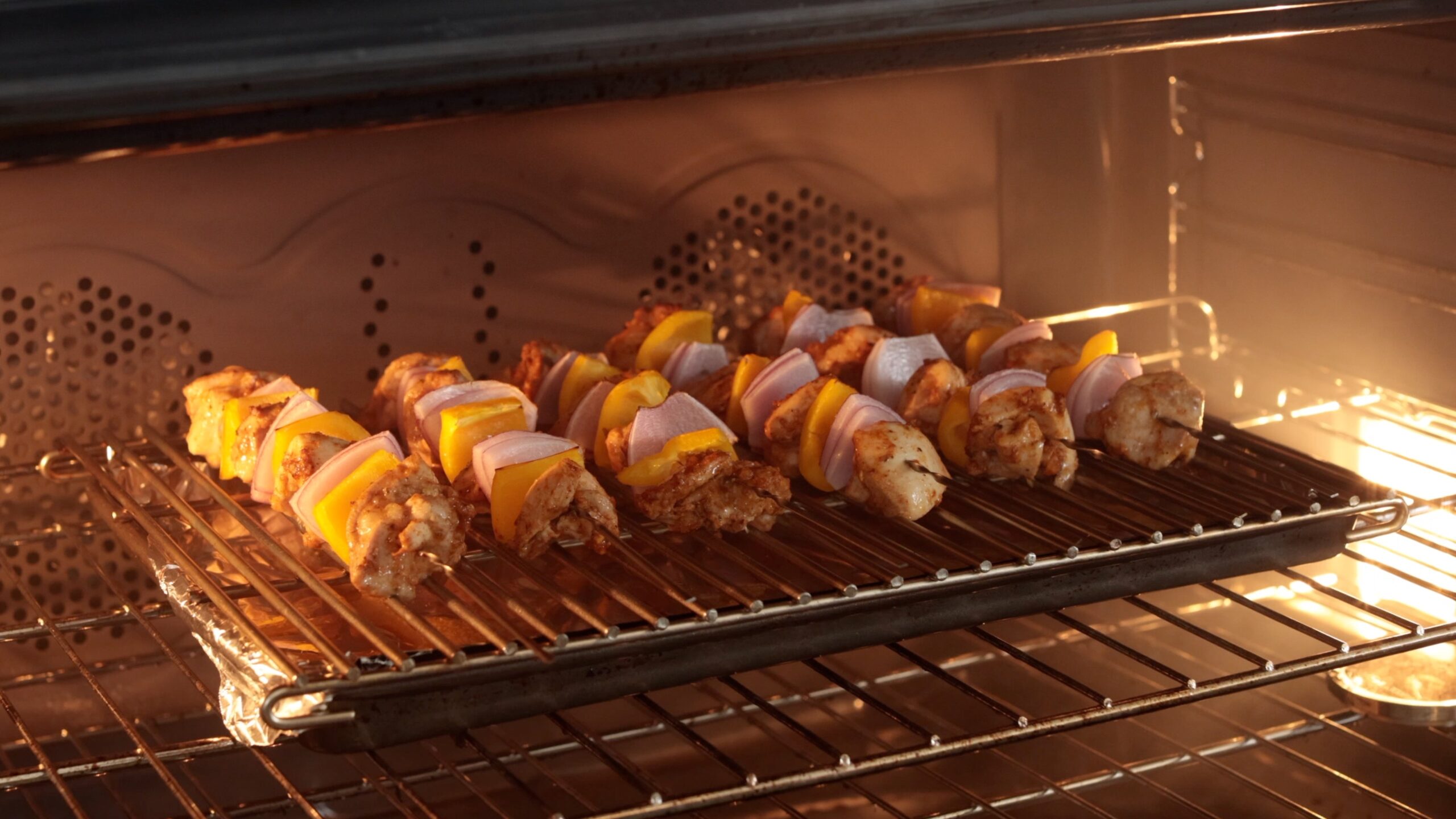 Chicken and vegetable skewers, with pieces of yellow bell pepper and onion, are arranged on a rack and roasting in an oven, illuminated by the oven light.