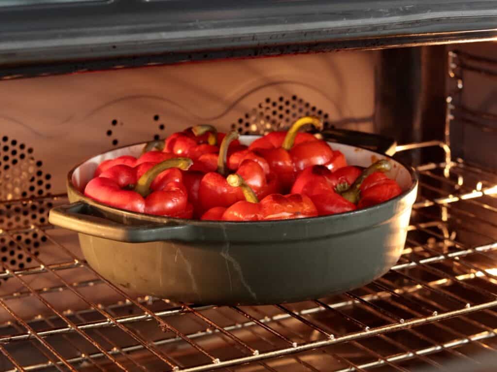 A green roasting pan filled with red bell peppers sits on an oven rack, roasting under the oven's light.
