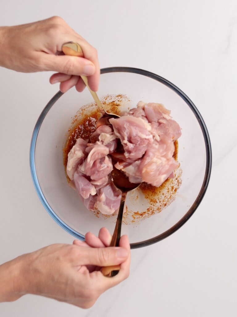 A person uses two spoons to mix raw, chopped chicken pieces with marinade in a clear glass bowl on a white surface.