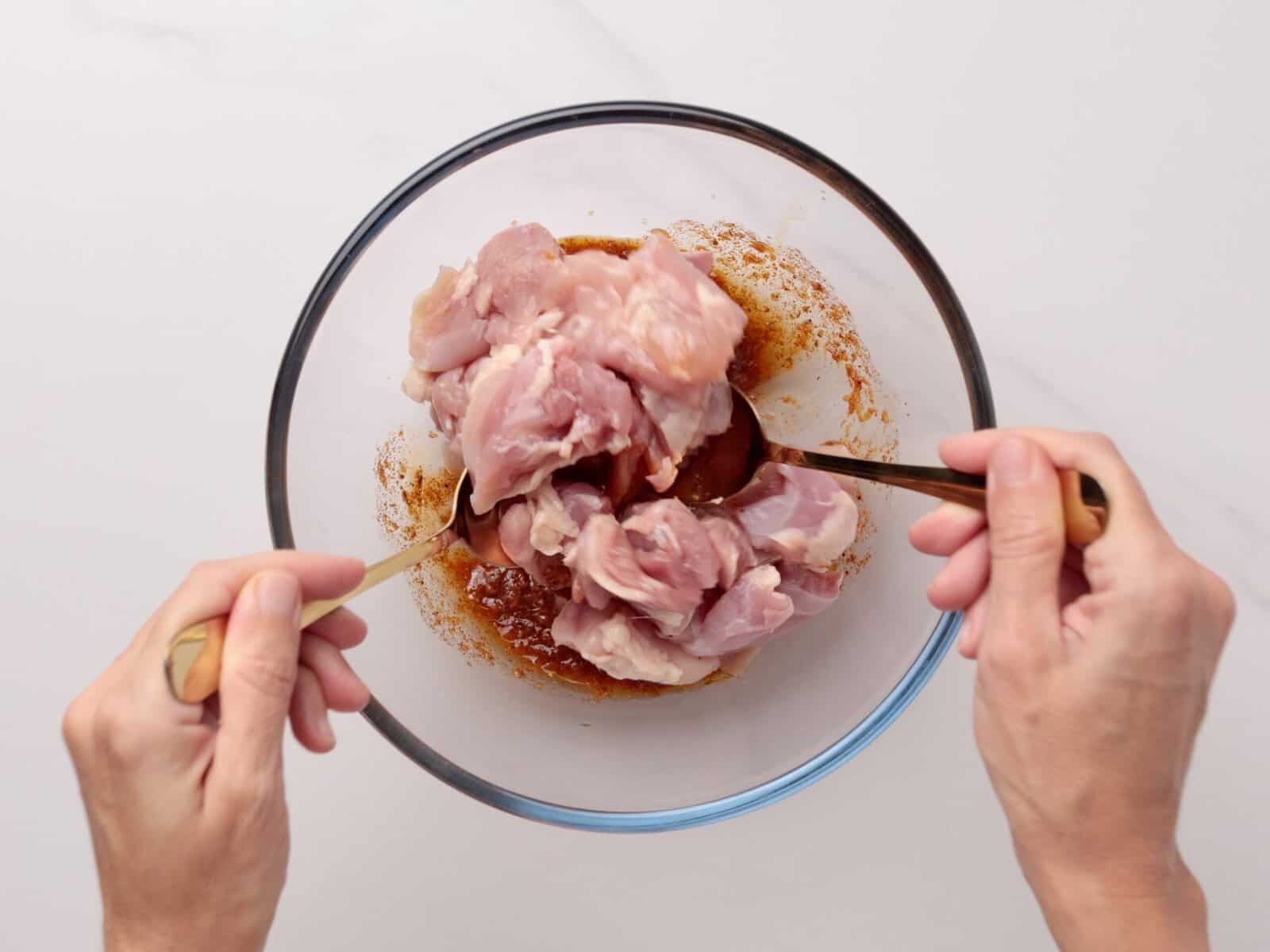 A person uses two forks to mix raw chicken pieces with marinade in a clear glass bowl on a white surface.
