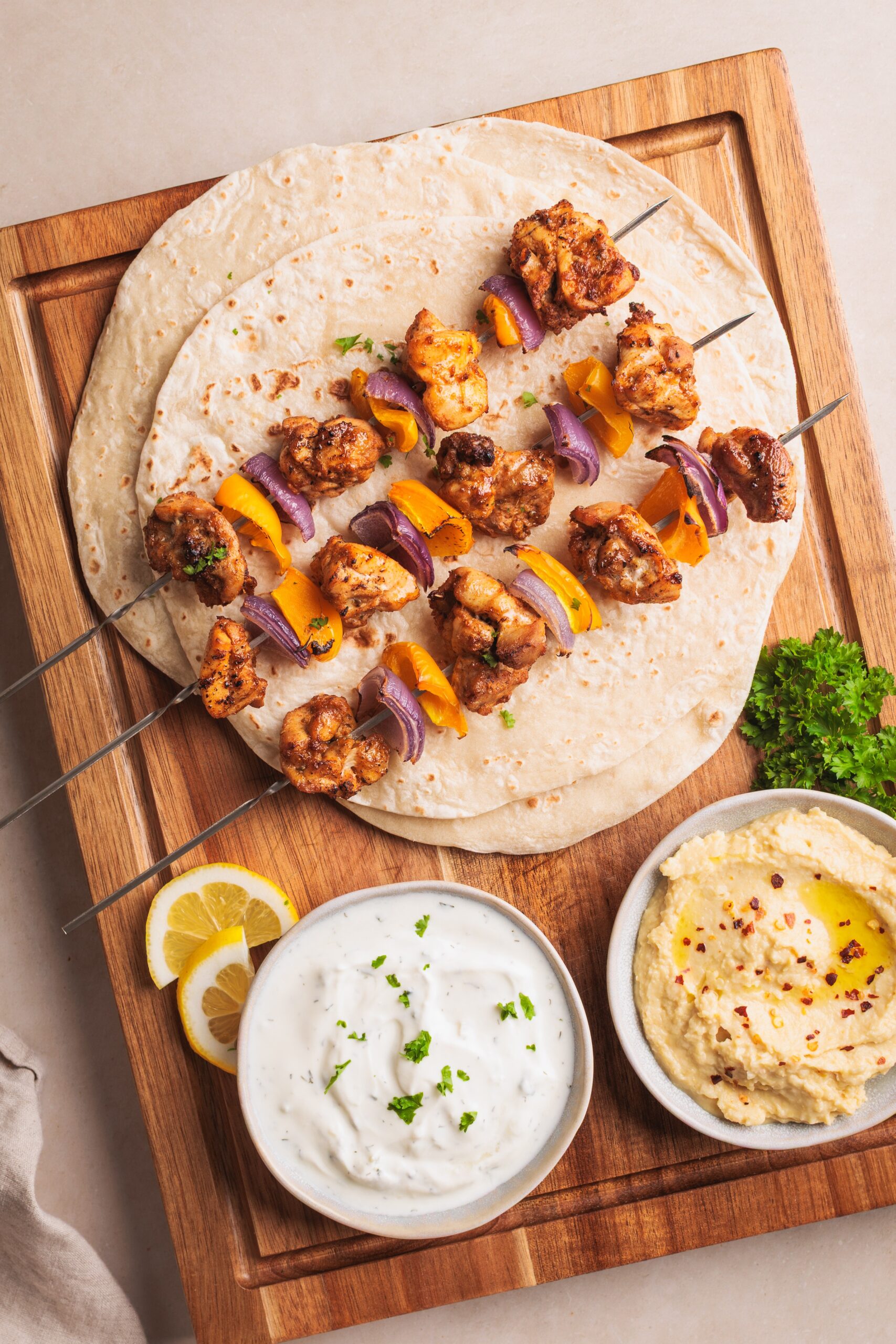 Two flatbreads with grilled vegetable and cauliflower skewers on top, served on a wooden board with bowls of white dipping sauce and hummus, lemon wedges, and parsley garnish.