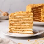 A slice of layered honey cake with cream filling sits on a white plate, with another piece of the same cake in the background. The cake has golden-brown layers and a crumb topping.