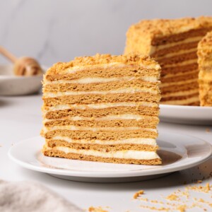 A slice of layered honey cake with cream filling sits on a white plate, with another piece of the same cake in the background. The cake has golden-brown layers and a crumb topping.