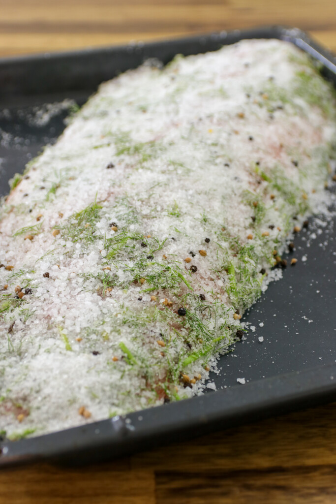 A large piece of fish covered in salt, dill, and pepper sits on a black baking tray, ready for curing or baking. The background is a wooden surface.