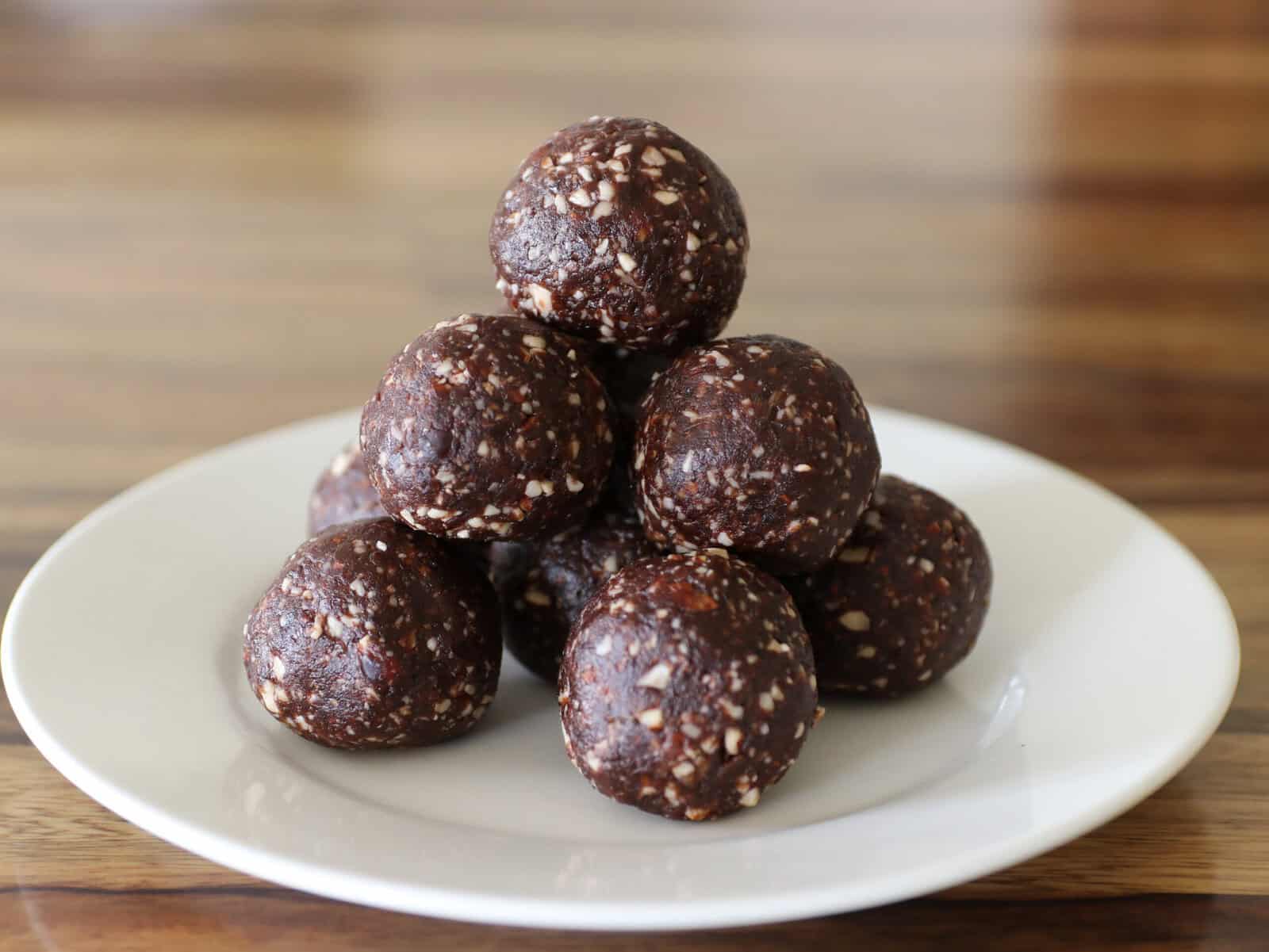 A white plate holds a pyramid of nine round, dark brown date energy balls with visible bits of nuts, set on a wooden table.