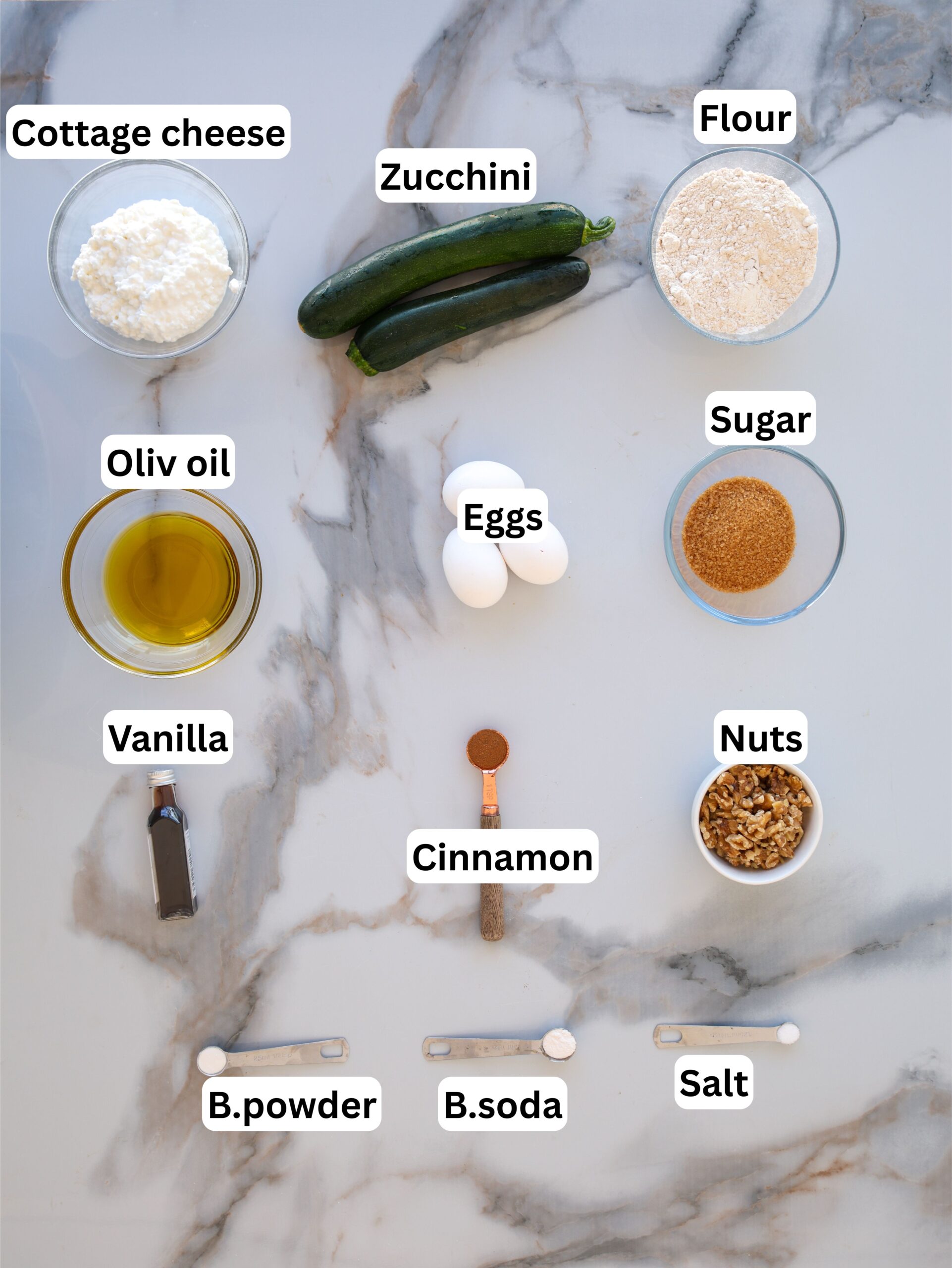 Overhead view of labeled baking ingredients on a marble surface, including cottage cheese, zucchini, flour, olive oil, eggs, sugar, vanilla, cinnamon, nuts, baking powder, baking soda, and salt.