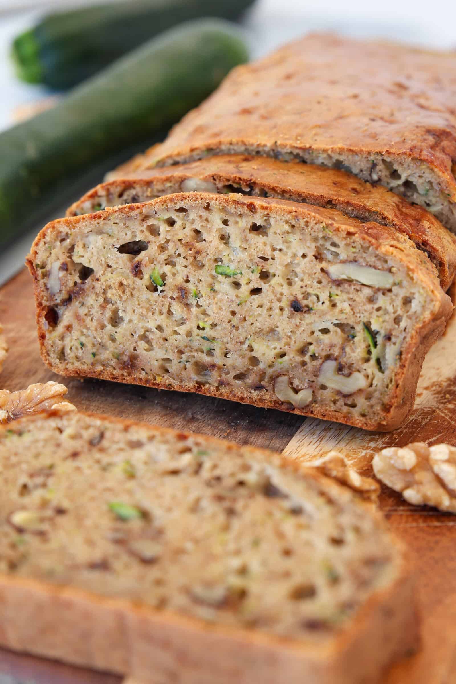 A close-up of a sliced cottage cheese zucchini bread with visible pieces of walnuts on a wooden board. Two zucchinis and walnut halves are in the background. The bread appears moist and speckled with green zucchini shreds.