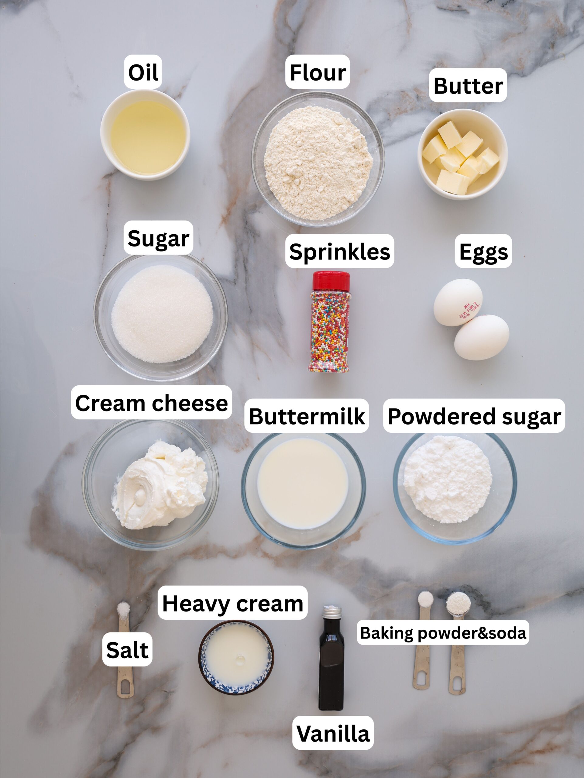 Various baking ingredients in bowls and cups arranged on a marble surface, including oil, flour, butter, eggs, sugar, sprinkles, cream cheese, buttermilk, powdered sugar, heavy cream, salt, vanilla, and baking powder & soda.