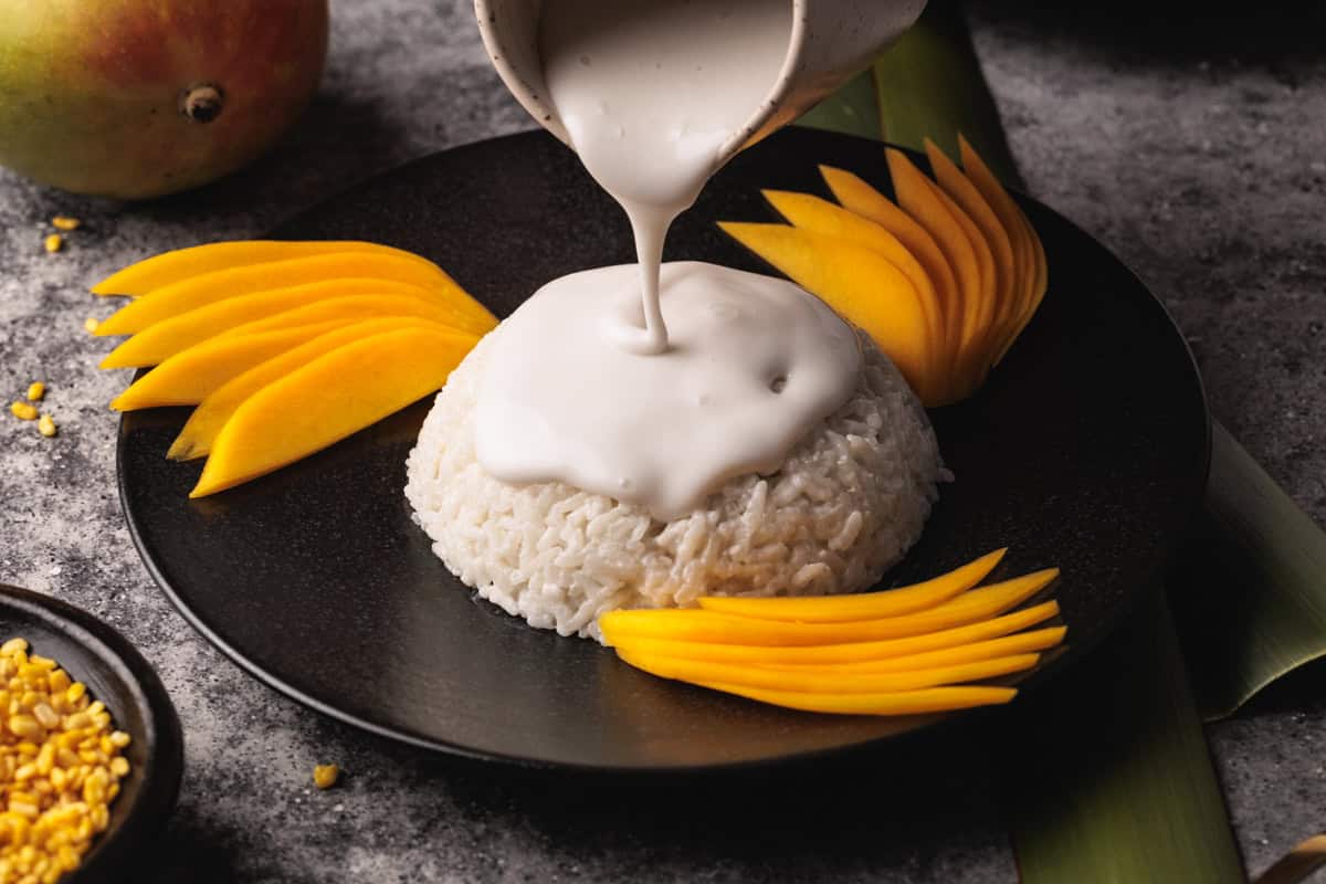 A serving of sticky rice on a black plate is topped with coconut sauce being poured from a small jug, surrounded by mango slices. A mango and a bowl of yellow beans are in the background.