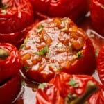 Close-up of stuffed red tomatoes baked in sauce, garnished with chopped herbs. The filling appears to be a tomato-based mixture with small vegetable pieces. The tomatoes look tender and juicy.