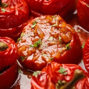 Close-up of stuffed red tomatoes baked in sauce, garnished with chopped herbs. The filling appears to be a tomato-based mixture with small vegetable pieces. The tomatoes look tender and juicy.