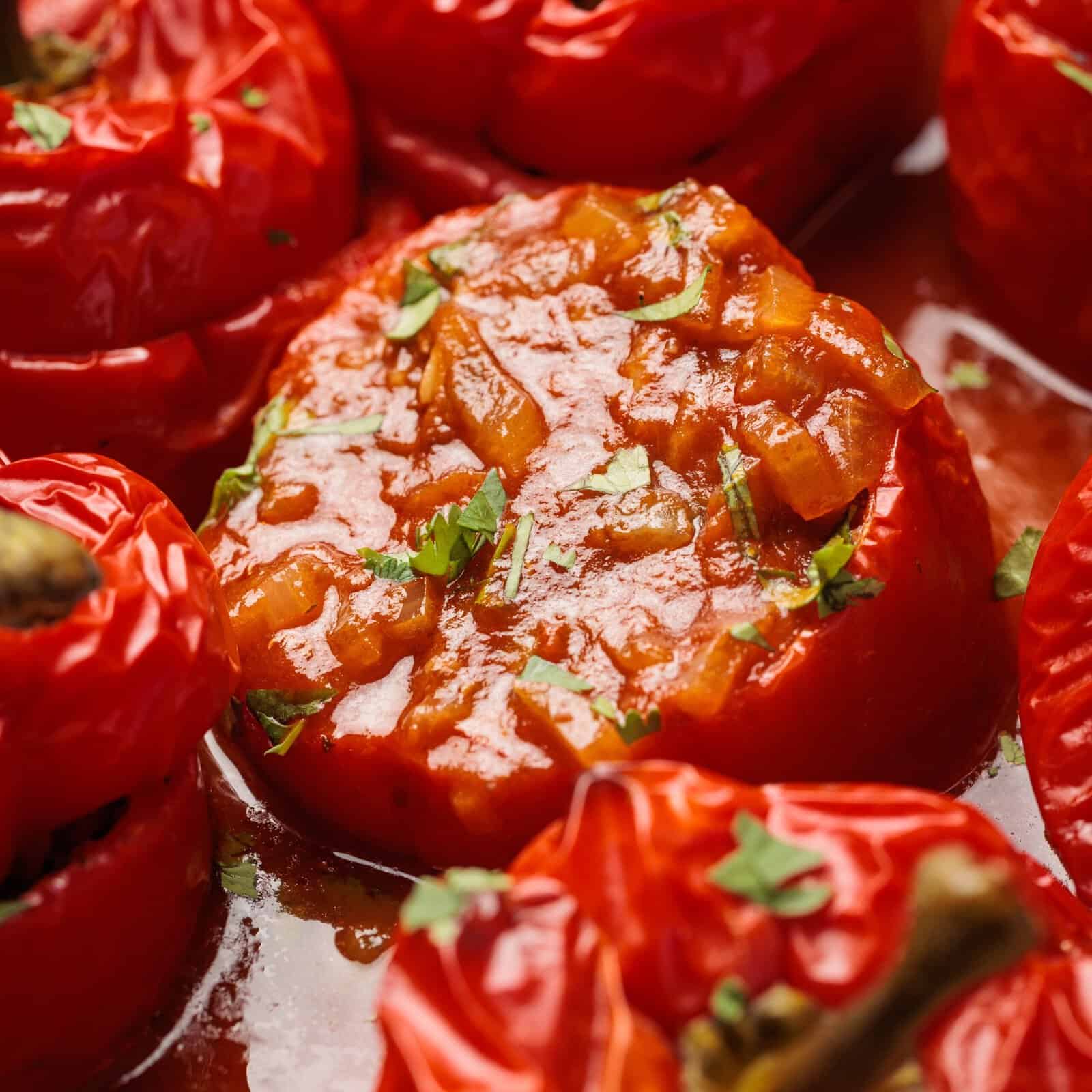Close-up of stuffed red tomatoes baked in sauce, garnished with chopped herbs. The filling appears to be a tomato-based mixture with small vegetable pieces. The tomatoes look tender and juicy.