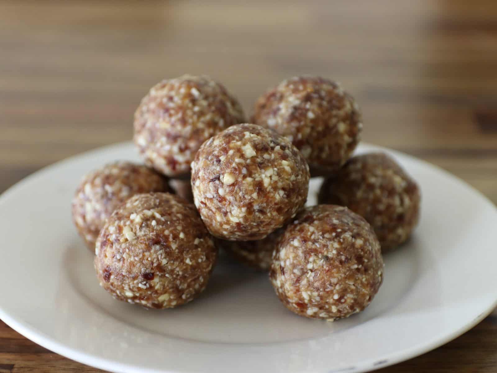 A white plate holds a pile of round, energy balls made with dates, visible pieces of nuts and seeds, set on a wooden surface.
