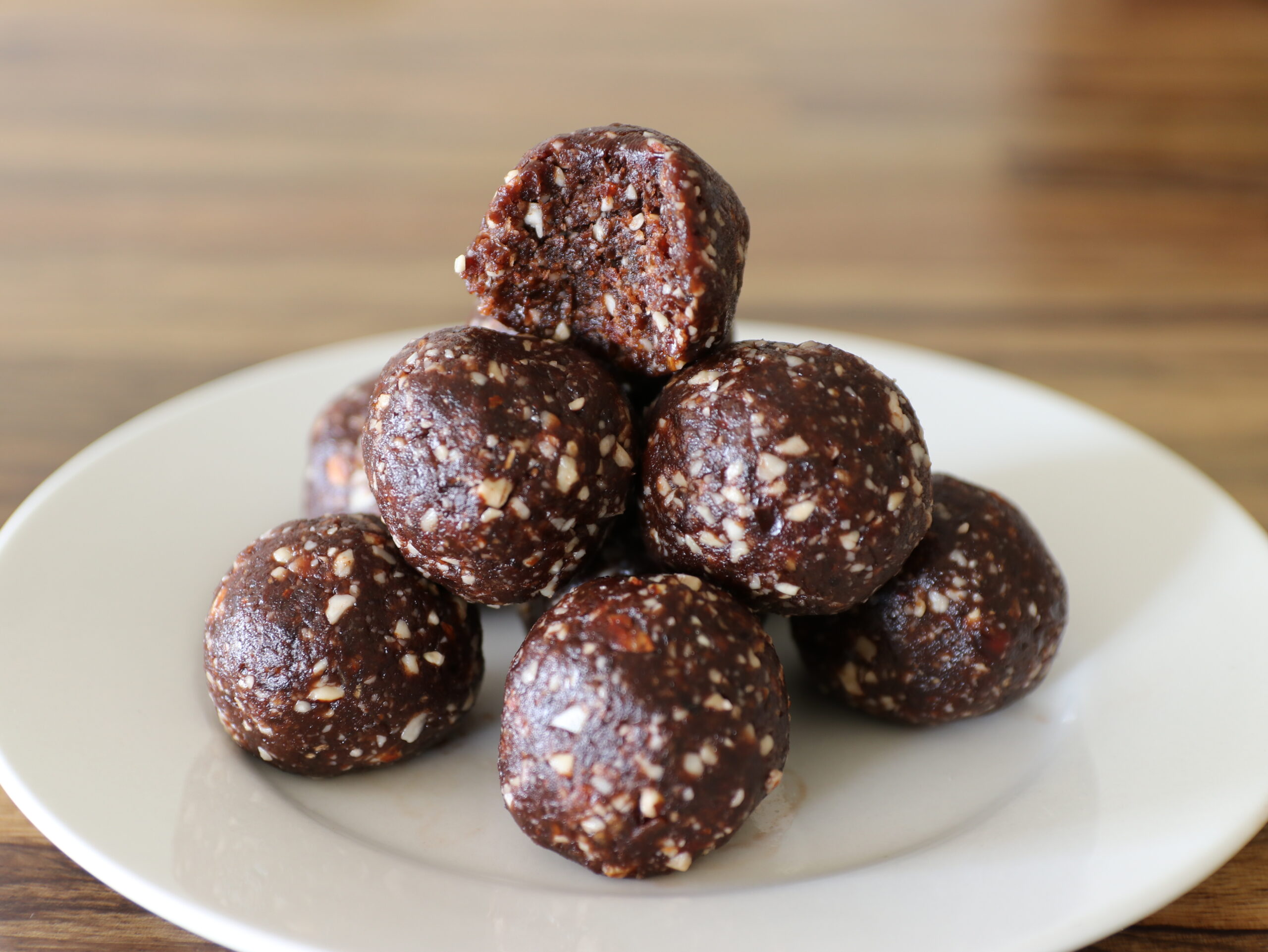 A stack of chocolate energy balls with visible nut pieces sits on a white plate, with the top ball showing a bite taken out. The background is blurred brown wood.
