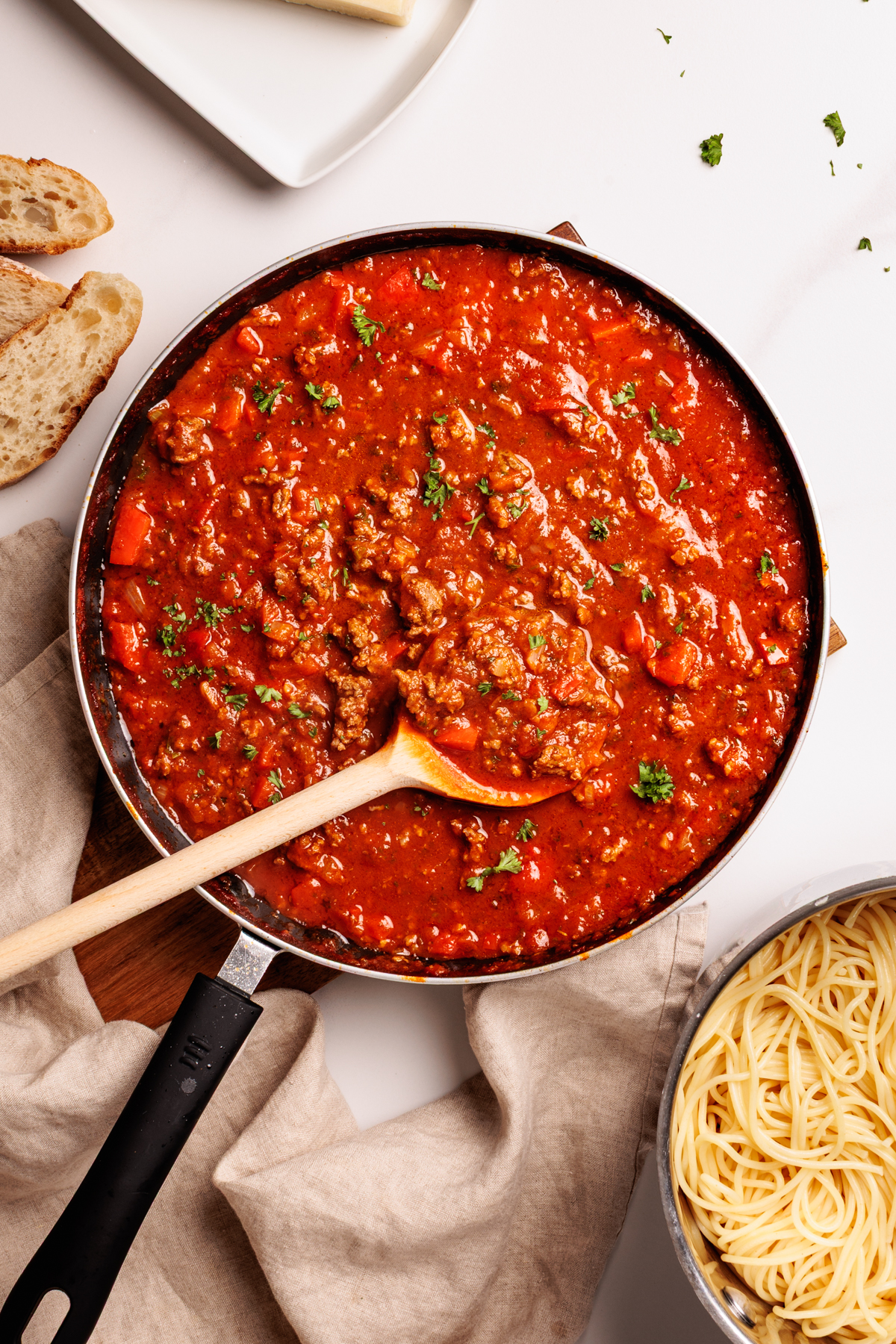 A skillet filled with chunky red meat sauce, garnished with parsley, sits on a cloth with a wooden spoon. Slices of bread, a plate, and a bowl of cooked spaghetti are nearby on a white surface.