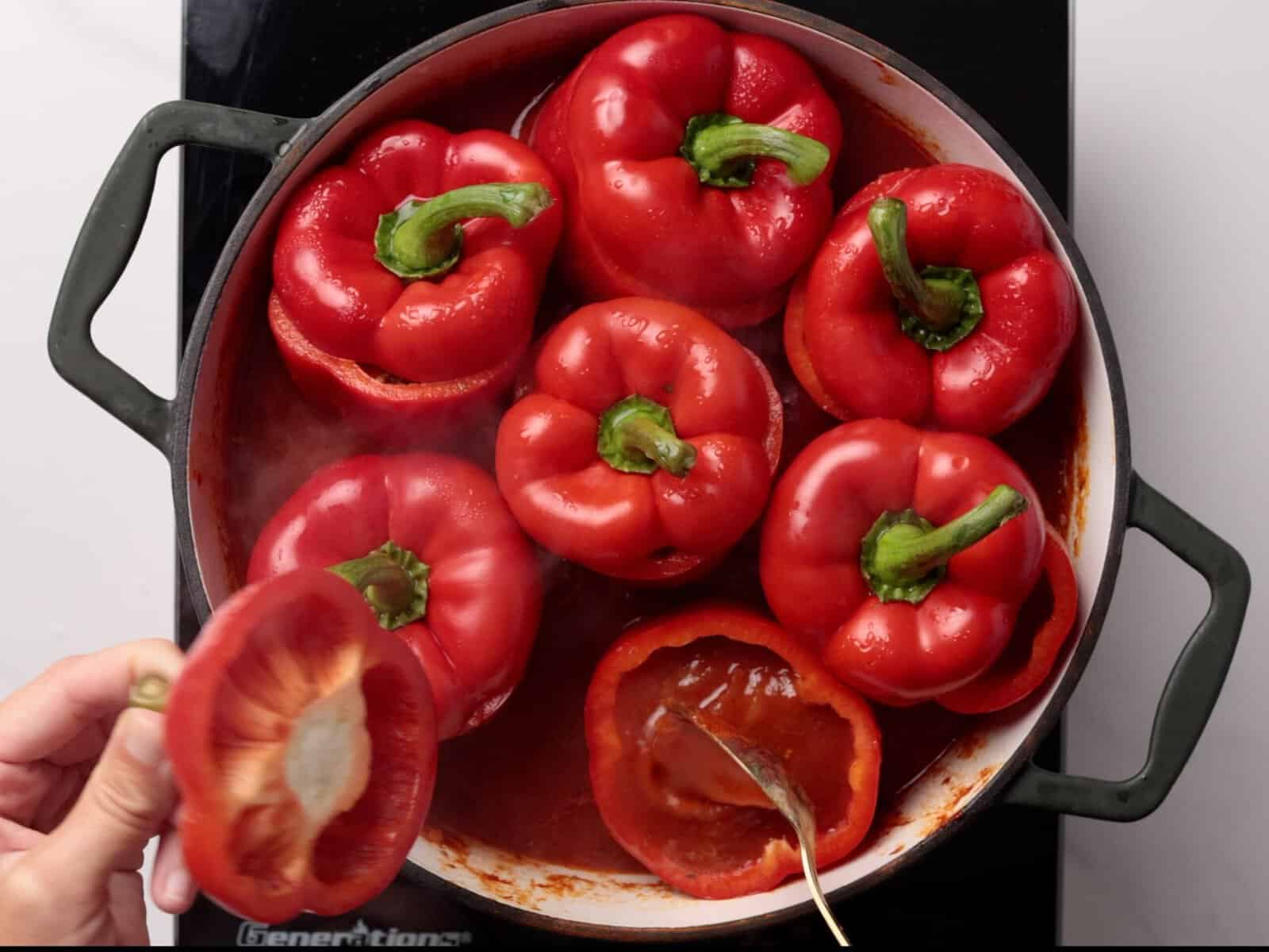 A hand holds the top of a red bell pepper above a pot filled with stuffed red bell peppers simmering in tomato sauce on a stovetop.
