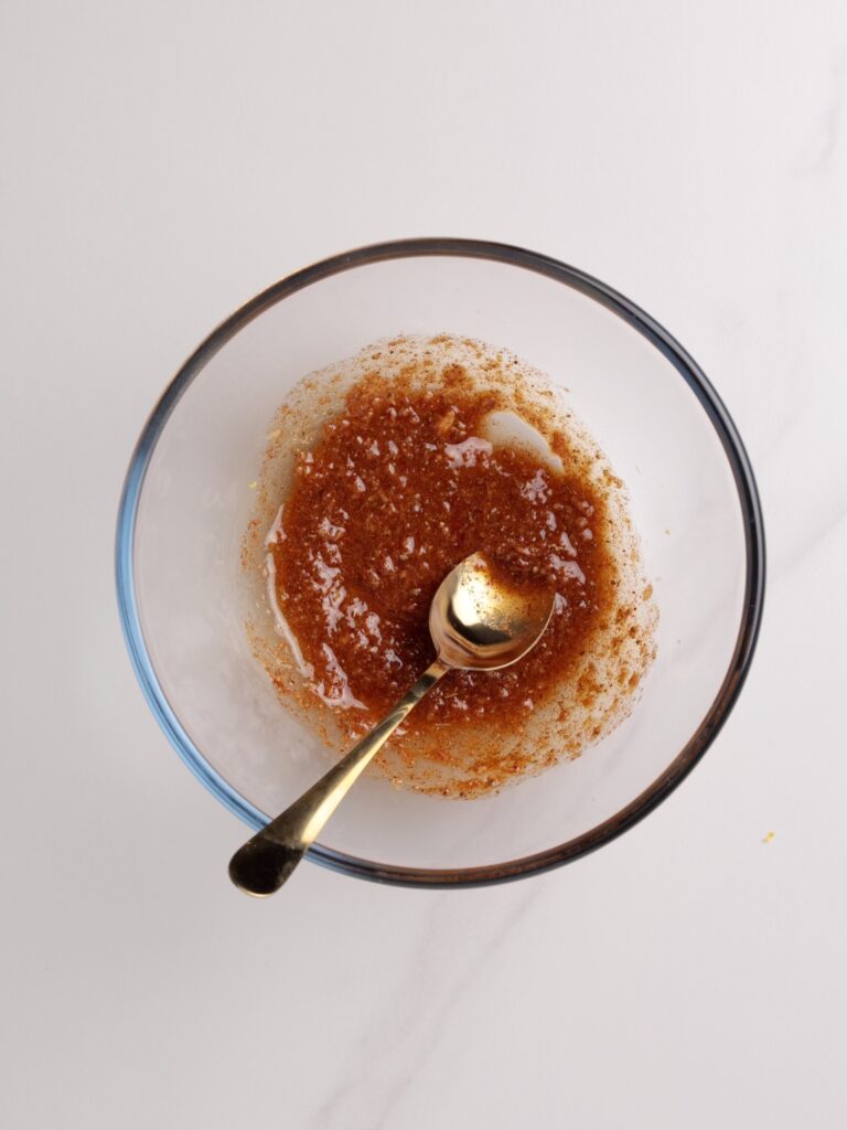 A glass bowl with a mixture of brown spices and liquid, being stirred with a silver spoon, sits on a white surface.