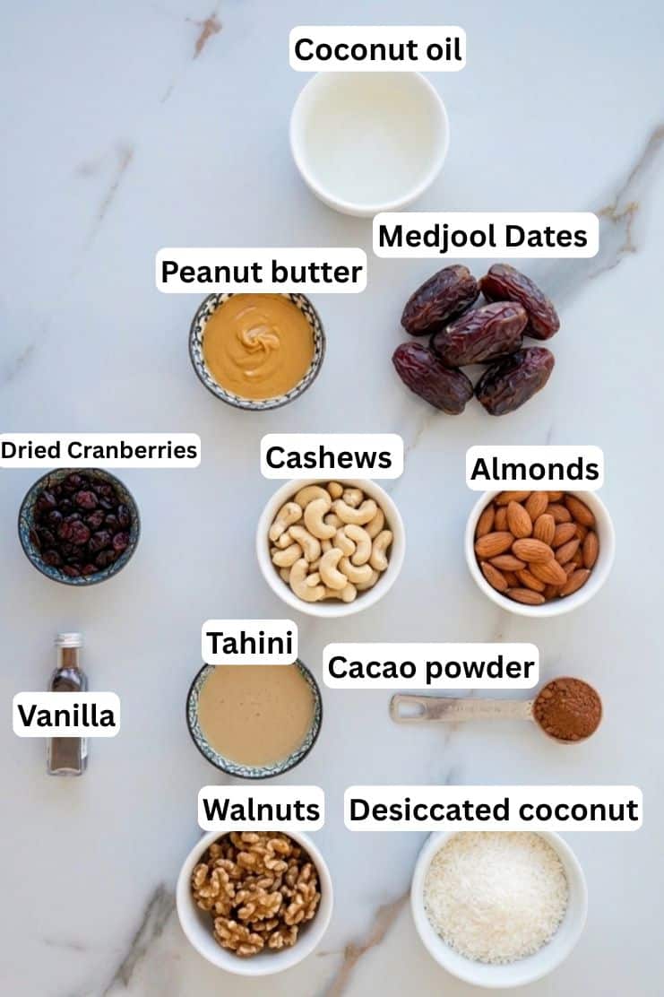 A flat lay of labeled bowls and containers holding coconut oil, Medjool dates, peanut butter, dried cranberries, cashews, almonds, tahini, cacao powder, vanilla, walnuts, and desiccated coconut on a white surface.