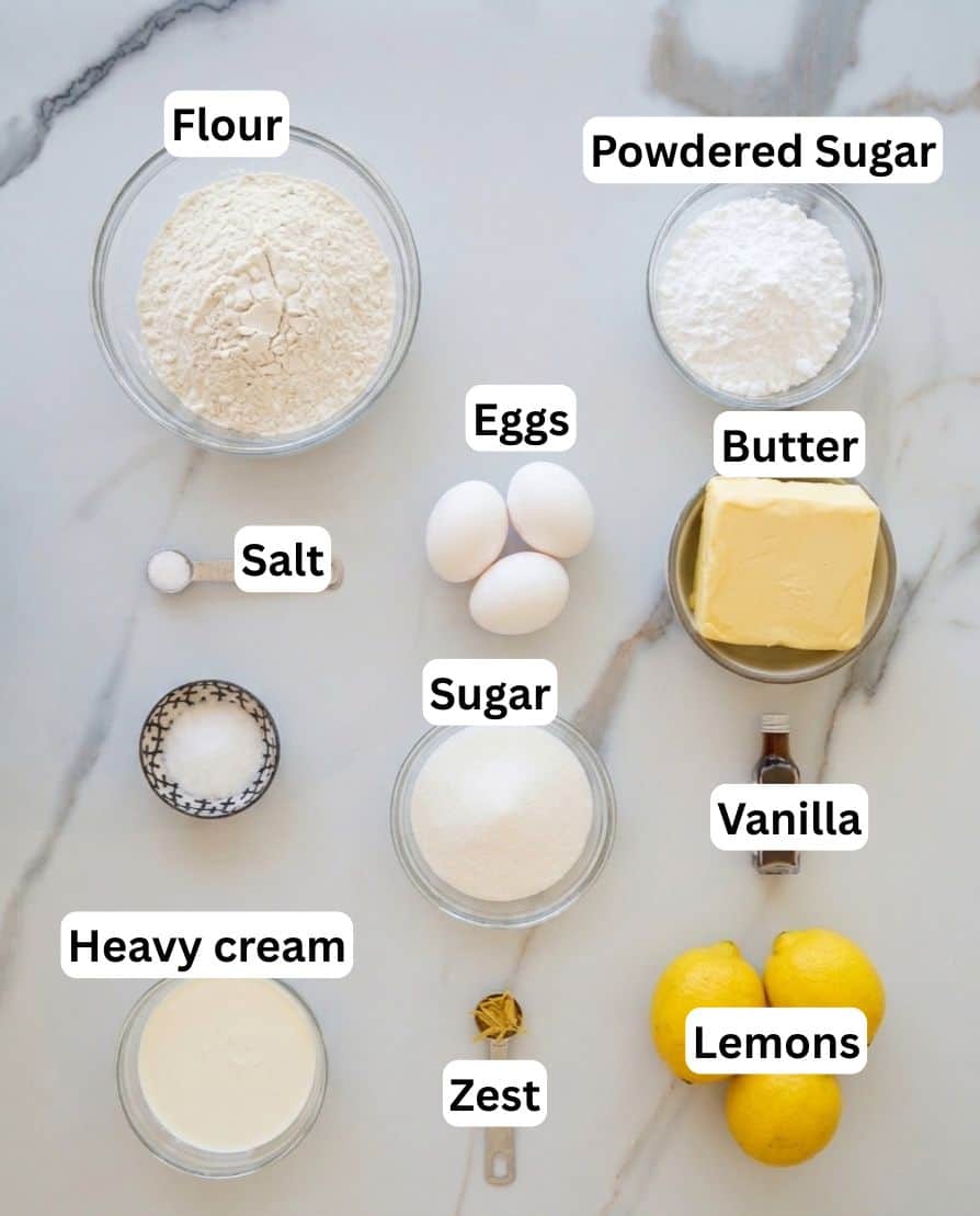Top-down view of labeled baking ingredients on a marble surface, including flour, powdered sugar, salt, eggs, butter, sugar, vanilla, heavy cream, lemons, and lemon zest, each in separate bowls or grouped.