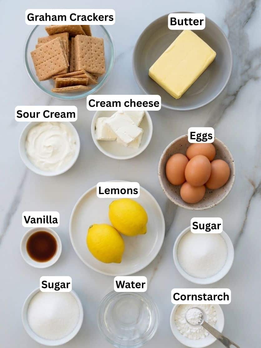 Labeled ingredients for cheesecake on a marble counter: graham crackers, butter, eggs, cream cheese, sour cream, lemons, water, vanilla, sugar (in two bowls), and cornstarch.