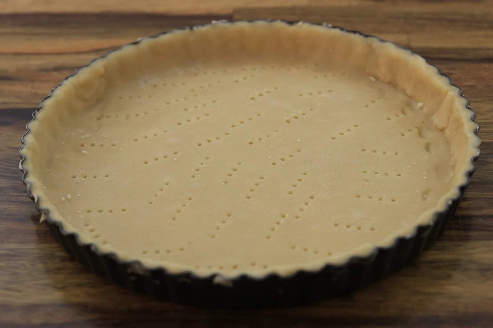 An unbaked tart crust with fork marks is pressed into a round tart pan and sits on a wooden surface.