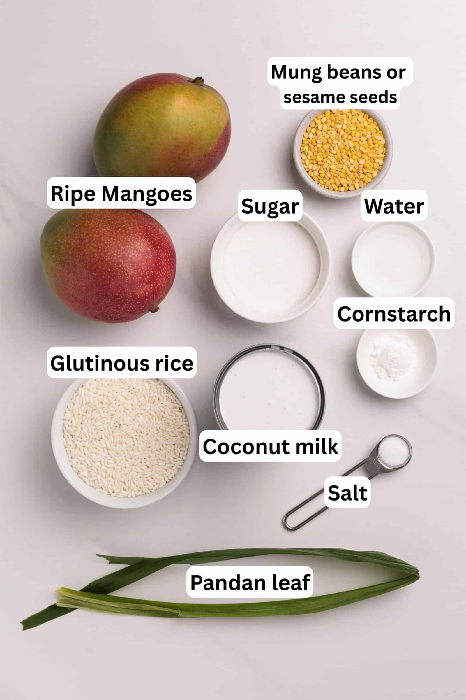 A flat lay of ingredients labeled for a dessert: ripe mangoes, mung beans or sesame seeds, sugar, water, cornstarch, glutinous rice, coconut milk, salt, and pandan leaf, all arranged on a white surface.