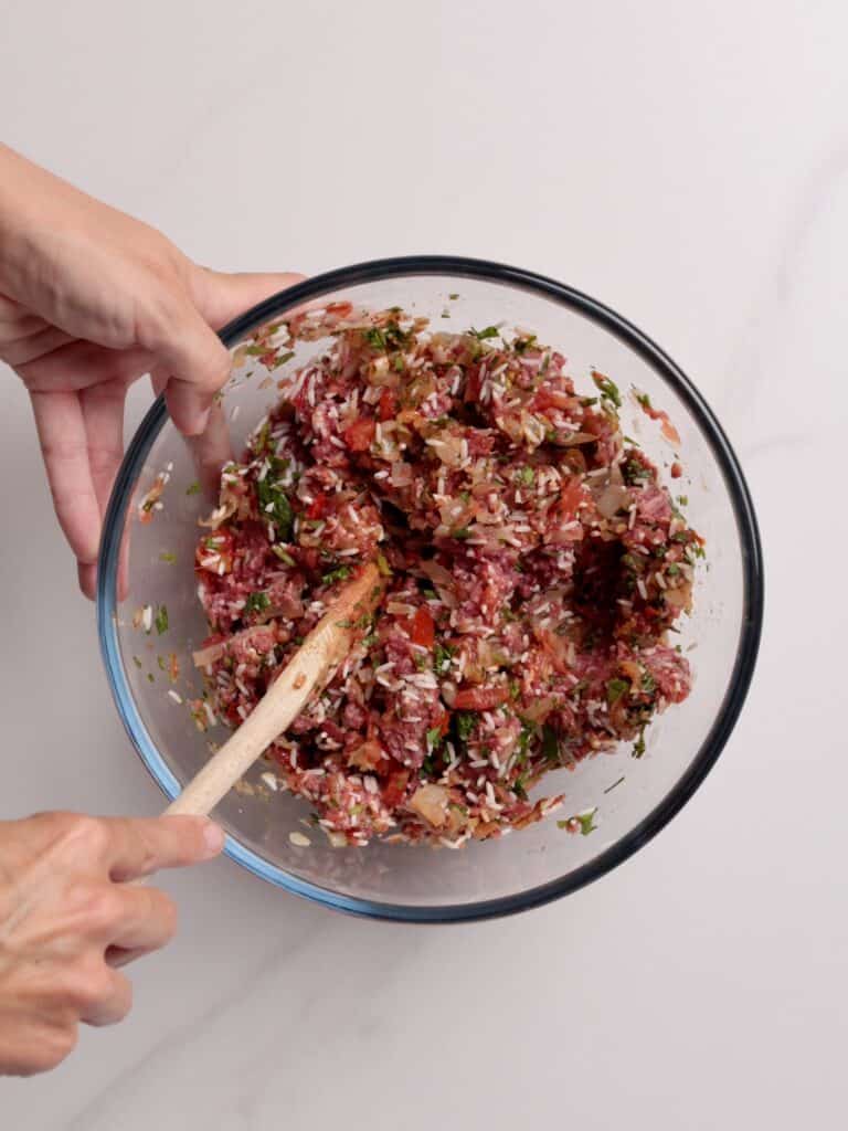 A person stirs a mixture of ground meat, herbs, and chopped vegetables for a stuffed-pepper recipe in a clear glass bowl with a wooden spoon on a white surface.