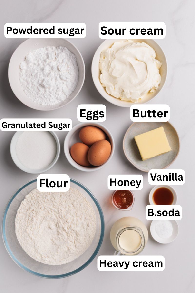 Top-down view of medovik ingredients in bowls on a white surface, including powdered sugar, sour cream, eggs, butter, granulated sugar, flour, honey, vanilla, baking soda, and heavy cream, each labeled with text.