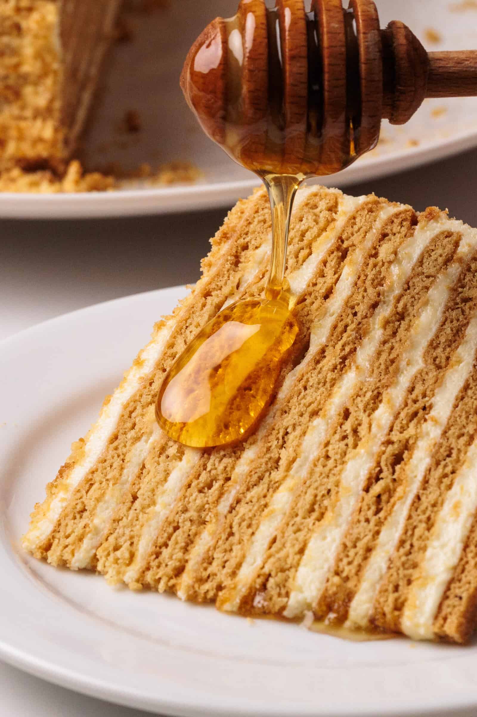 A slice of layered Russian honey cake on a white plate with honey being drizzled over it from a honey dipper.