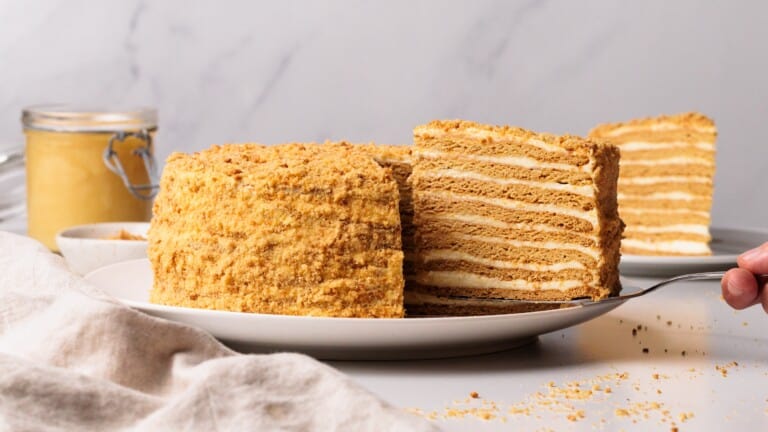 A layered Russian honey cake with golden crumbs on the outside sits on a plate. A slice is being served, revealing creamy layers inside. A jar and bowl are in the background on a light surface.