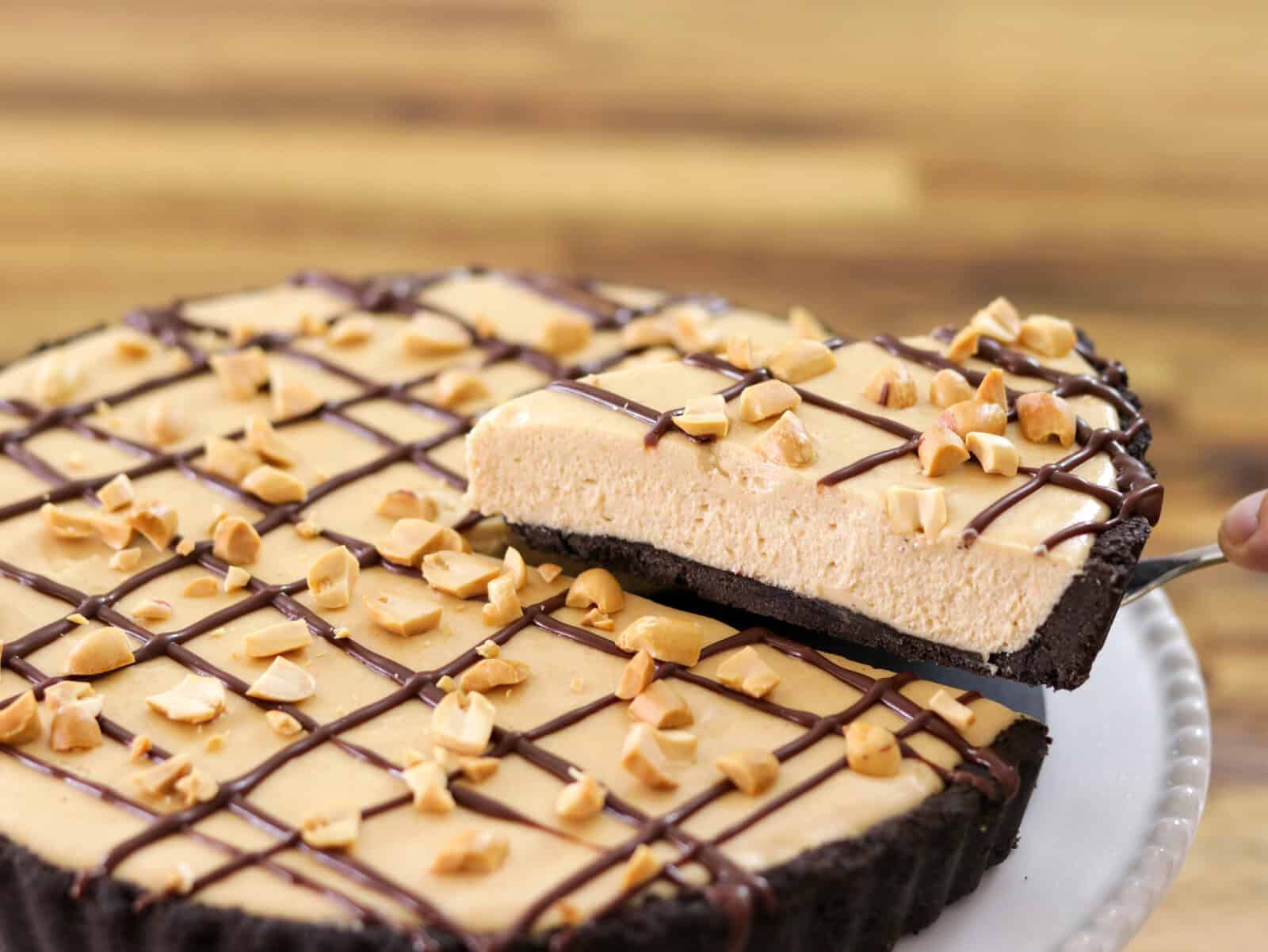A creamy peanut butter pie with a chocolate crust is topped with chopped peanuts and a drizzle of chocolate, with one slice being lifted from the pie on a server.