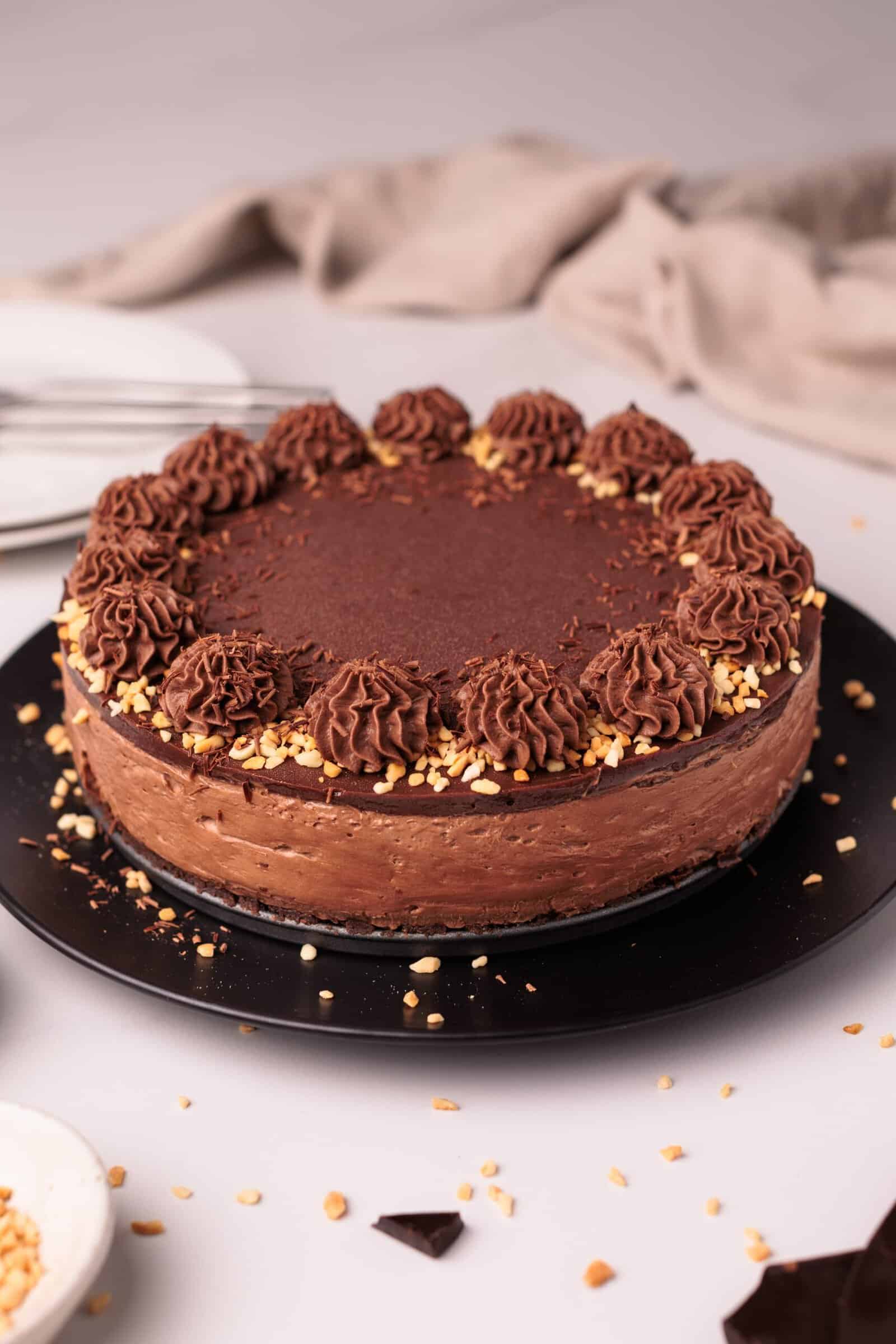 A round Nutella cheesecake on a black plate, decorated with piped chocolate frosting and sprinkled chopped nuts around the edge, sits on a light surface with a beige cloth and empty plates in the background.