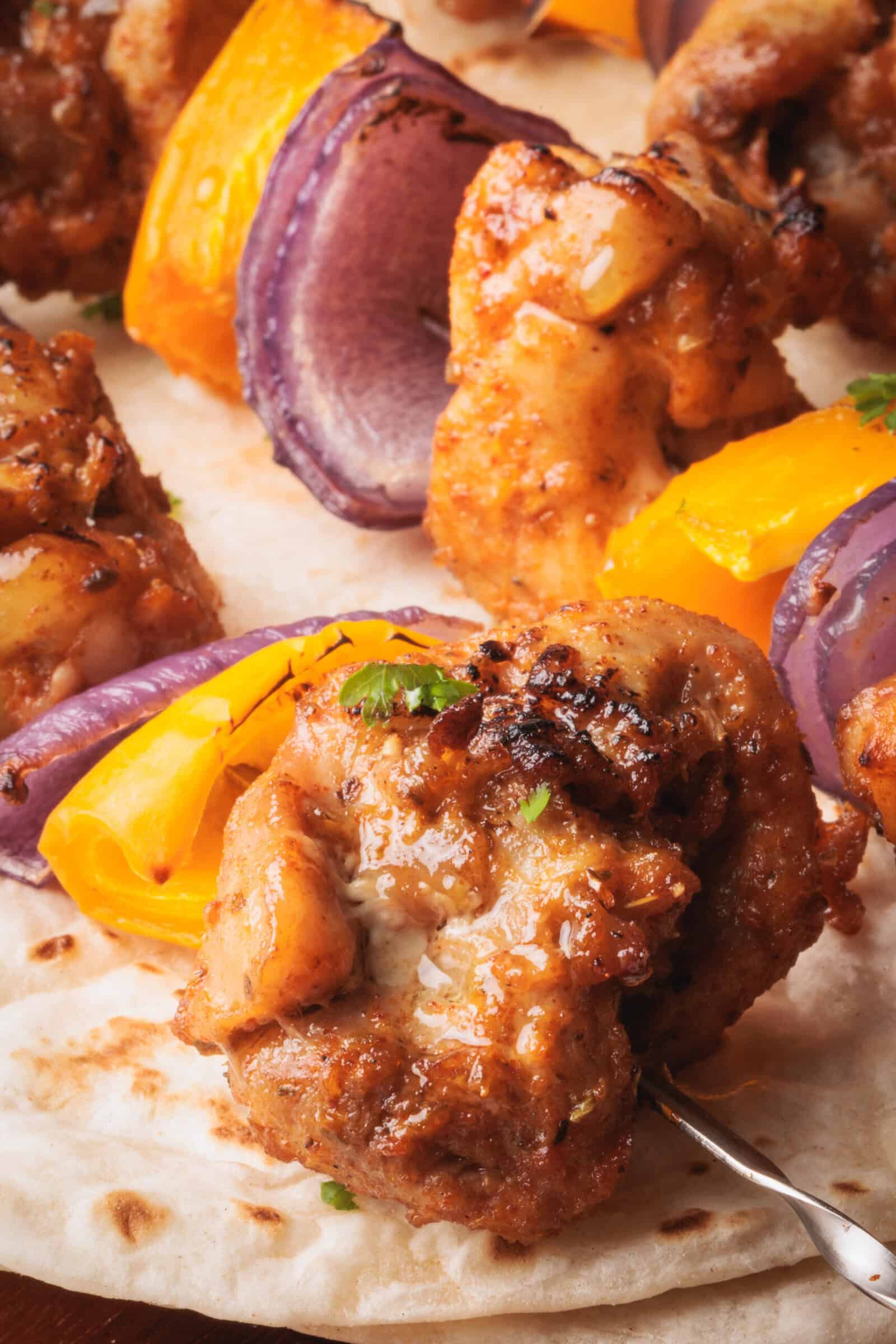 A close-up of oven-baked chicken skewers with slices of yellow bell pepper and red onion, served on flatbread. The chicken is browned and garnished with herbs.