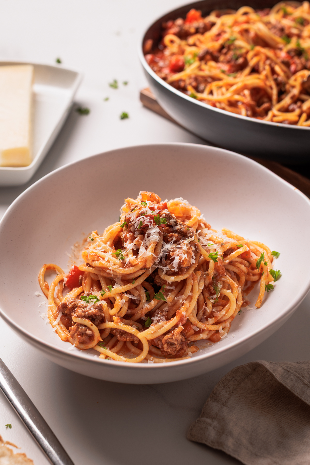 A bowl of spaghetti topped with meat sauce and sprinkled with grated cheese and parsley, with a pan of more pasta and a block of cheese in the background.