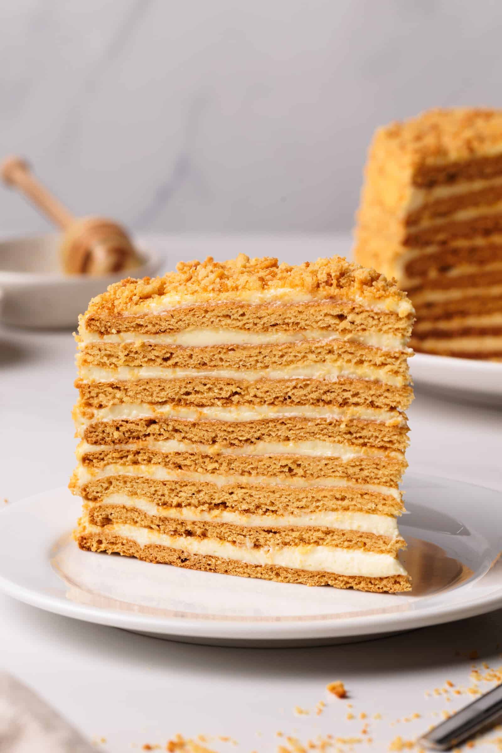 A slice of layered Russian honey cake (medovik) with alternating golden cake and cream layers sits on a white plate, with crumbs scattered around and another slice in the background.