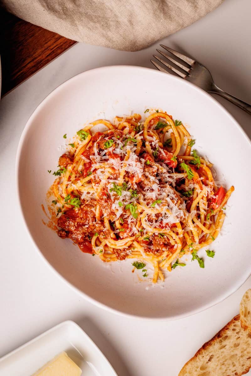 A white plate of spaghetti and meat sauce, grated cheese, and chopped parsley sits on a table next to a fork, bread, and a block of cheese.