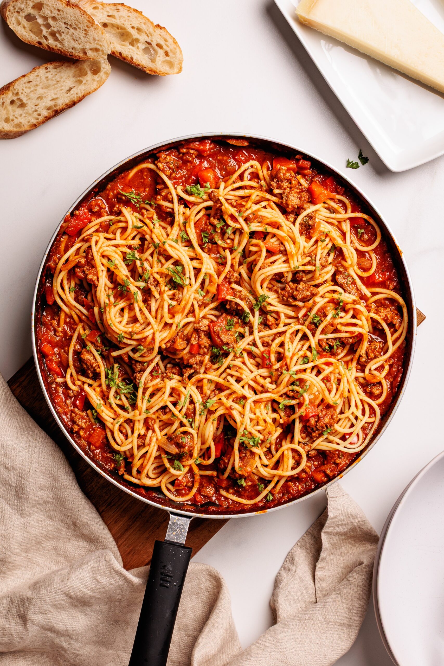A skillet filled with spaghetti mixed in a chunky tomato and ground meat sauce, garnished with herbs. Slices of bread and a block of cheese are nearby on a white surface.