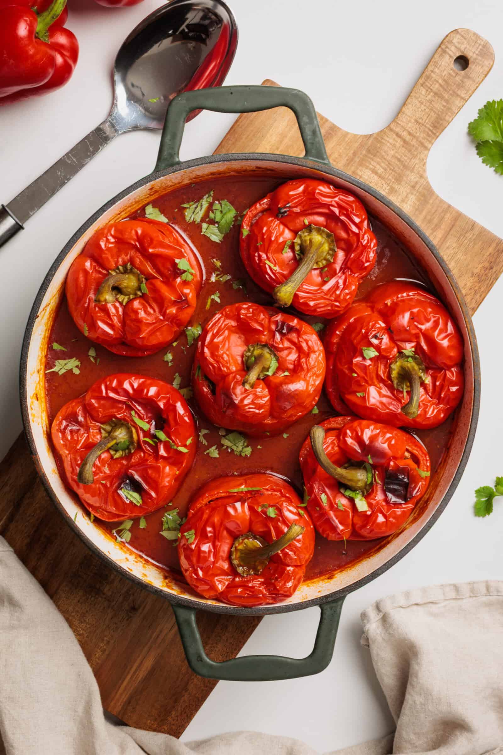 A round baking dish filled with eight stuffed red bell peppers in tomato sauce, garnished with fresh herbs, sits on a wooden board with a spoon, red pepper, and a beige napkin nearby.