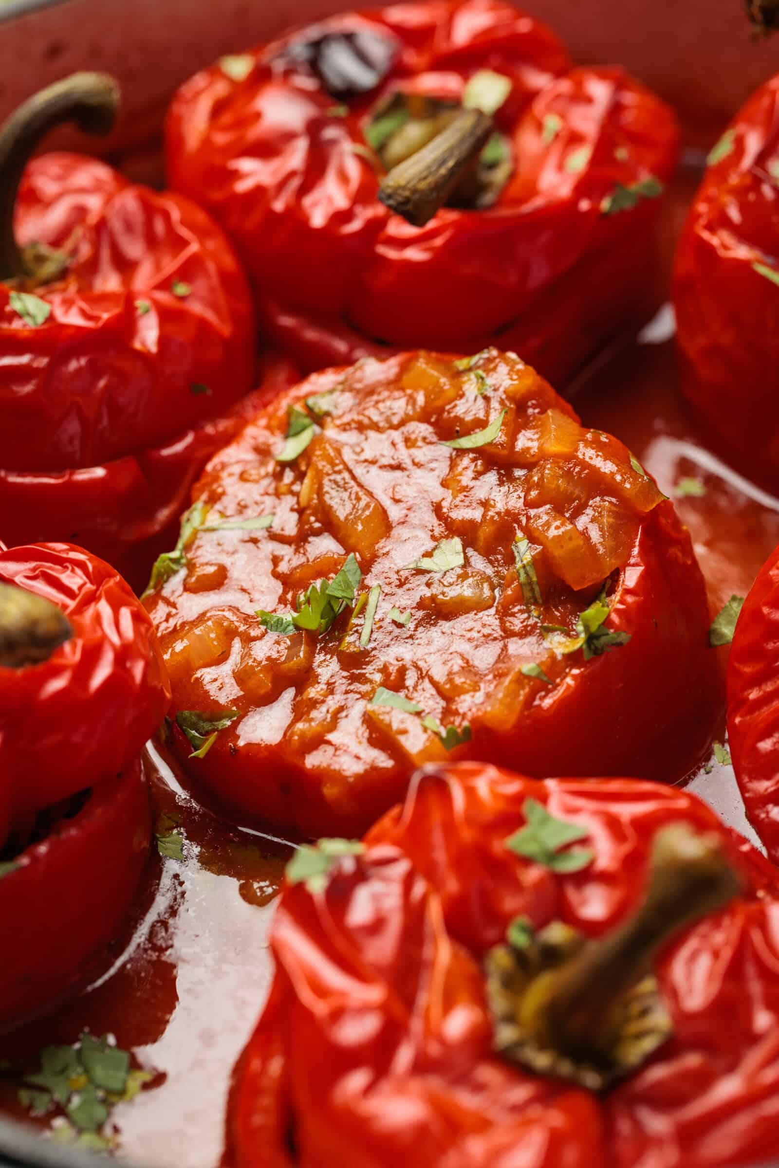 Close-up of roasted red bell peppers stuffed with a tomato-based mixture and garnished with chopped herbs, with some peppers showing their stems and shiny, baked skin.