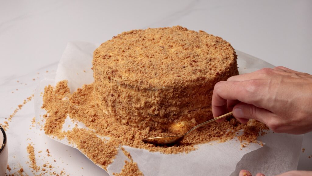 A hand uses a spoon to press crumb coating onto the side of a round cake, which is sitting on parchment paper and surrounded by loose crumbs.