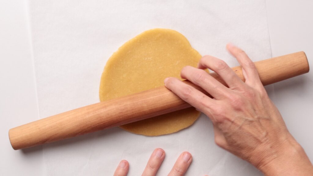 A person uses a wooden rolling pin to flatten a round piece of dough on a sheet of parchment paper.