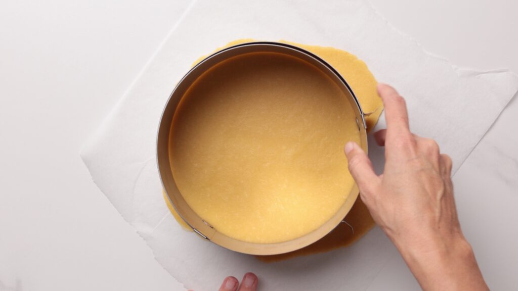 A hand places a round baking tin over rolled out dough on parchment paper, preparing to cut or shape the dough to fit the tin.