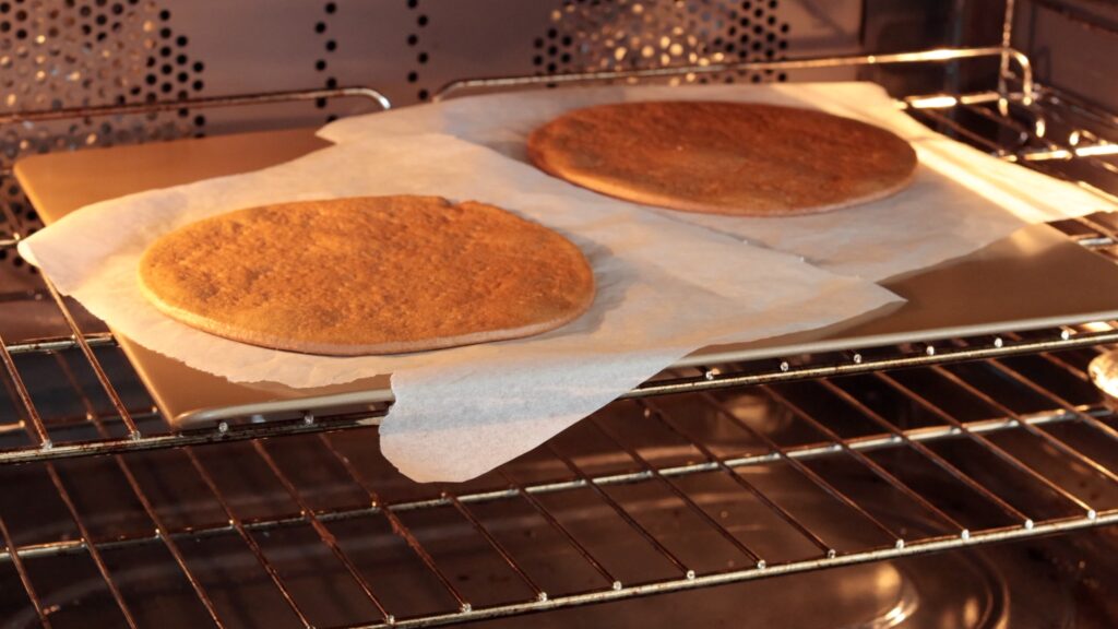 Two large, round cookies or flatbreads baking on parchment paper atop a baking tray inside an oven, with one appearing slightly darker than the other.
