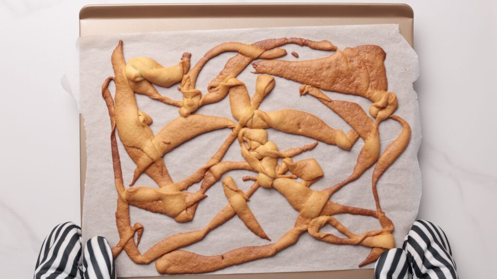 A baking tray with a sheet of parchment paper holds an unevenly spread, overbaked cookie dough, creating an abstract, tangled shape. Gloved hands hold the tray over a white surface.
