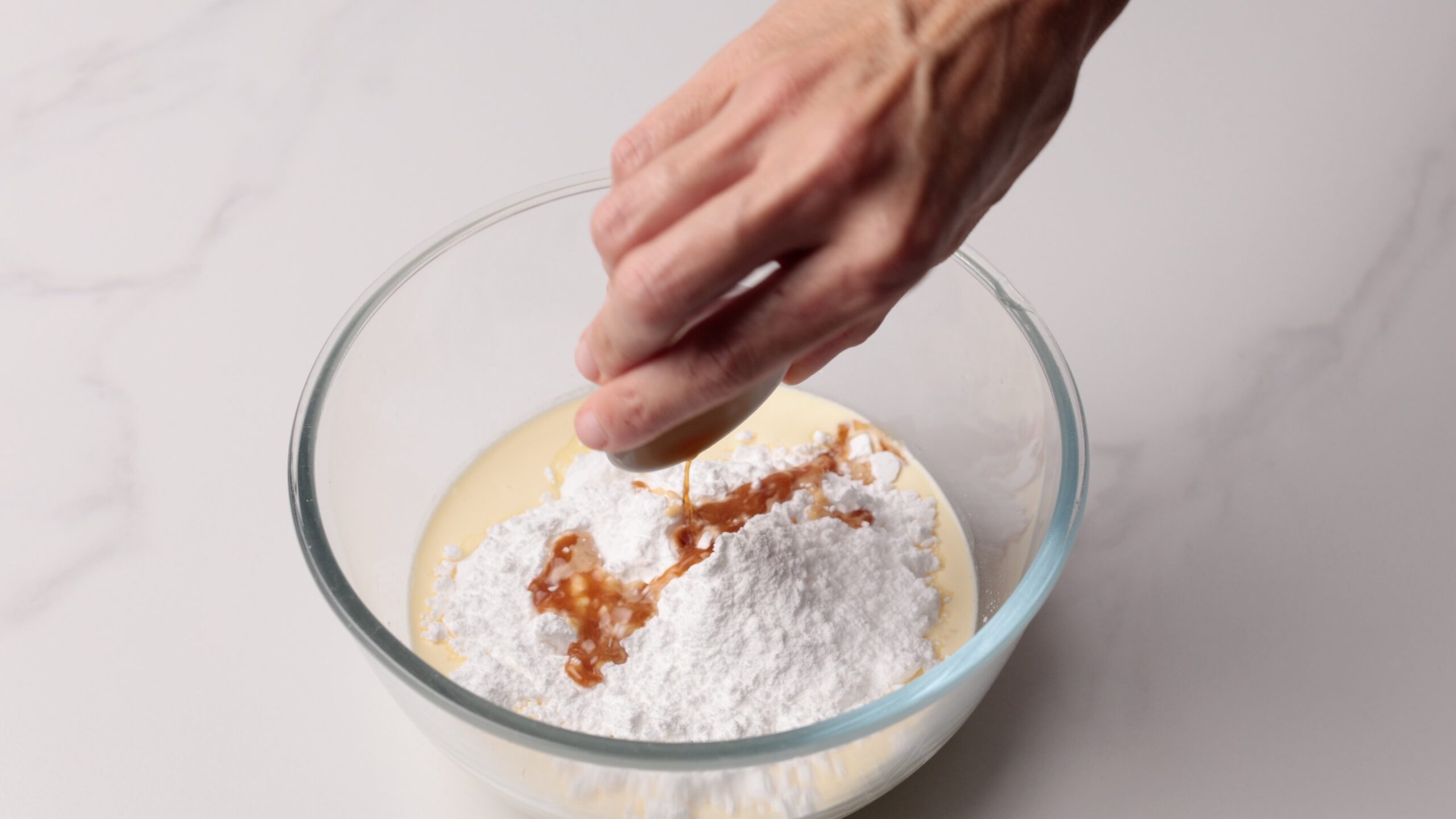 A hand pours liquid, likely vanilla extract, into a glass bowl containing powdered sugar and a creamy mixture, preparing ingredients for baking on a white surface.