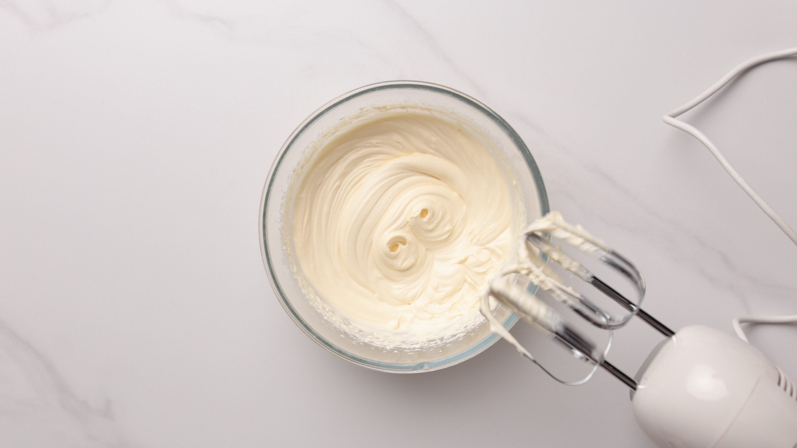 A glass bowl filled with creamy medovik filling mixture sits on a white surface. An electric hand mixer with metal beaters is partially visible on the right, coated with the same mixture.