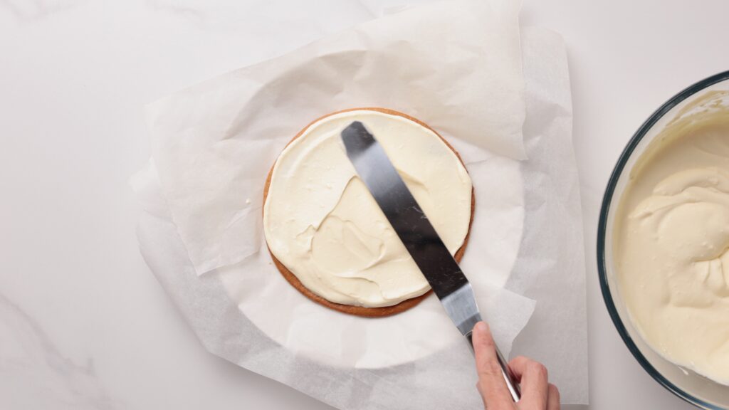 A hand uses an icing spatula to spread white frosting onto a round cake placed on parchment paper; a bowl of frosting is visible beside the cake.